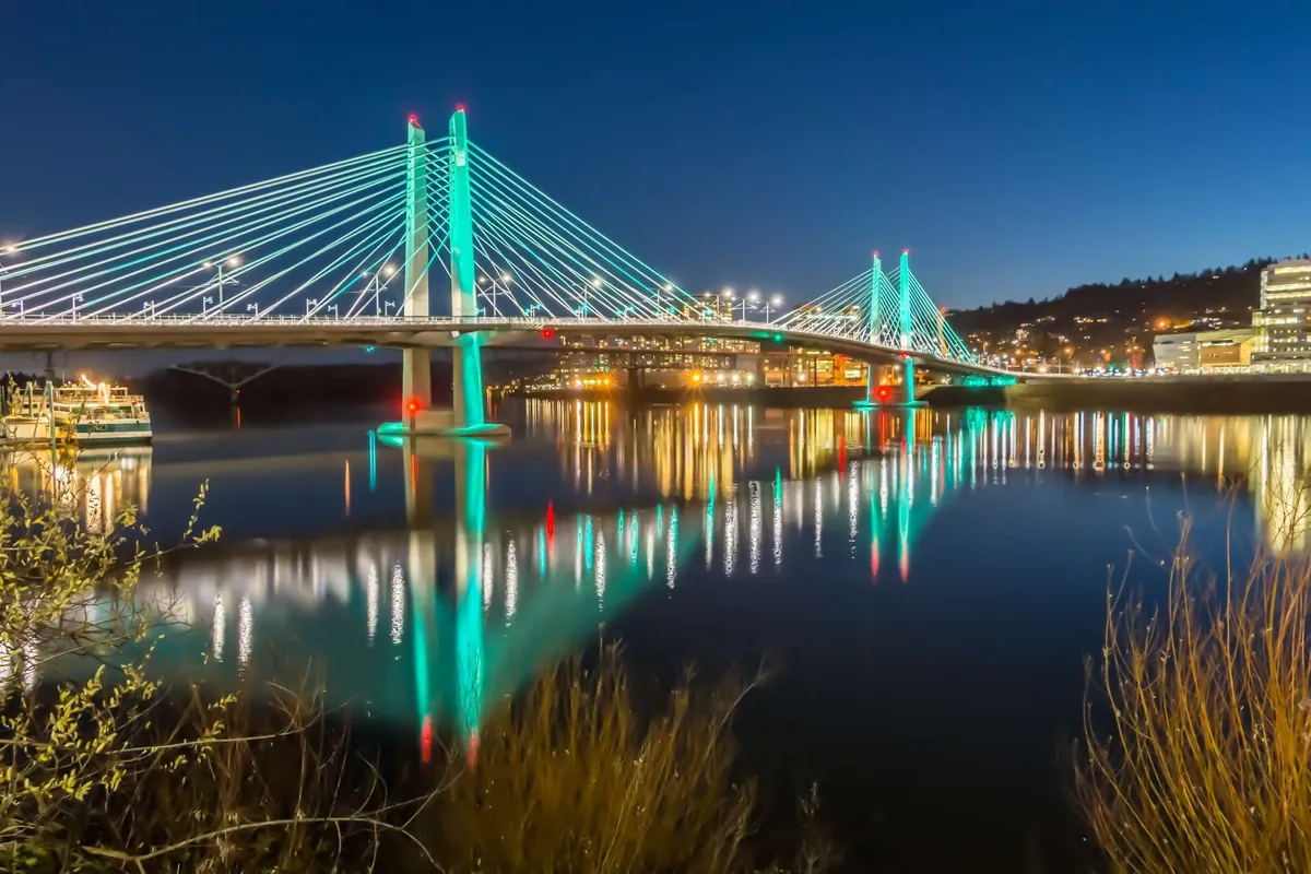 A scenic exterior shot featuring an illuminated bridge reflected in calm water, with the city lights creating a tranquil yet lively evening ambiance. The image evokes the spirit of local exploration and riverside strolls.