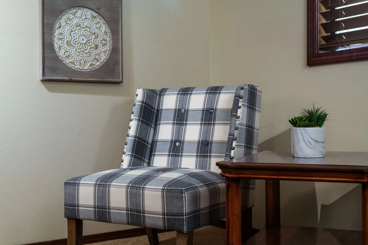 A cozy seating area featuring a plaid chair and wooden side table, adorned with decorative wall art and a small potted plant. The area feels inviting and is positioned for quiet reading, casual conversation, or a relaxing break between activities.