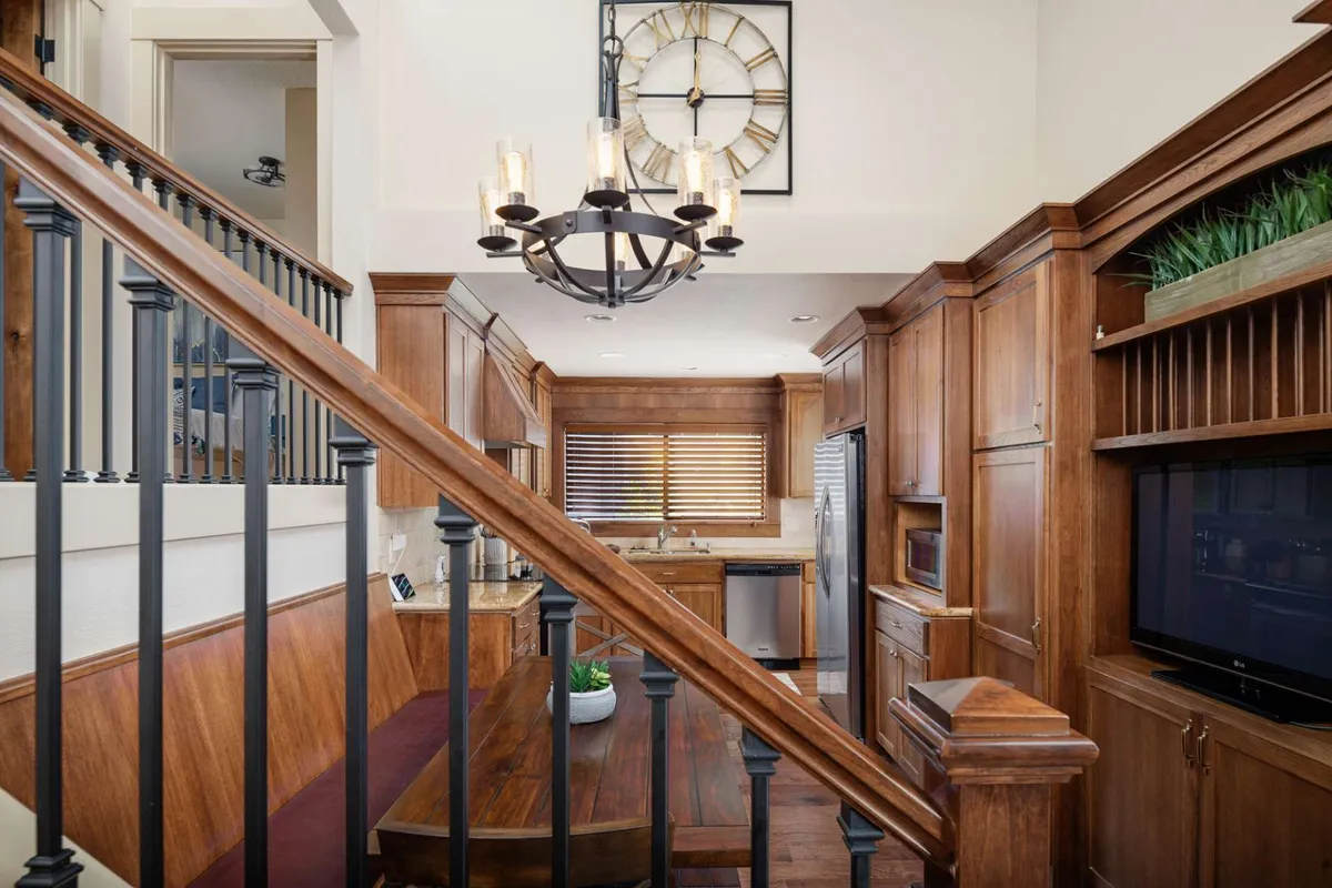 A warmly lit dining space featuring a prominent chandelier above a spacious wooden dining table, surrounded by wooden cabinetry and a visible wall clock. The table is set as the focal point for gatherings, while the open layout hints at kitchen proximity and social interaction.