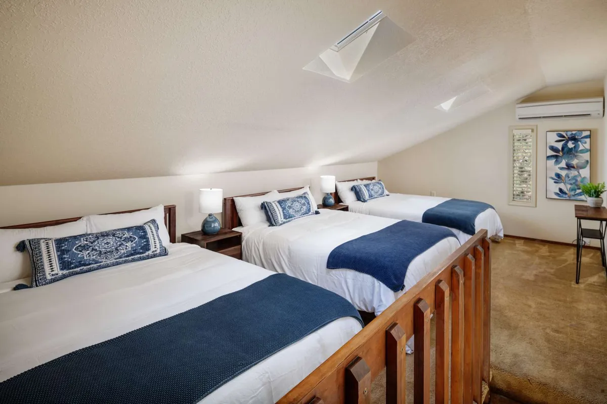 A spacious bedroom featuring three beds with decorative pillows, a small table with a plant, and inviting lighting. The room is designed for comfort and togetherness, making it ideal for groups or families to share.