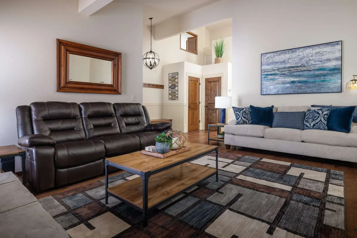A spacious living room featuring a brown leather sofa and a light-colored sofa arranged around a wooden coffee table, complemented by a decorative painting and a modern light fixture. The room is bathed in natural light, with plush seating and inviting decor encouraging guests to relax and connect.