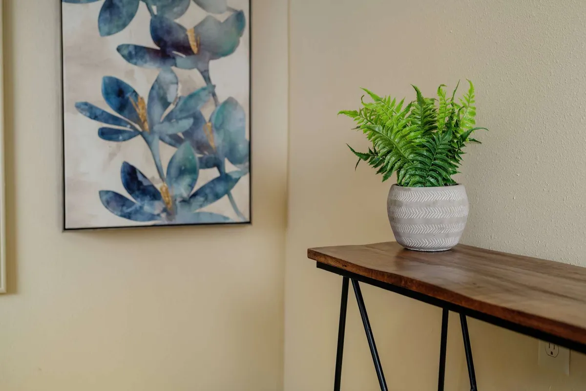 A tranquil corner featuring a wooden console table adorned with a potted fern and floral artwork, set against a softly lit wall. The space feels open and inviting, suggesting a pleasant transition area near the entrance or a spot to pause after coming indoors.