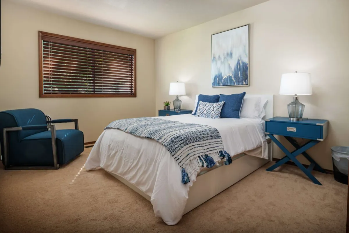 A serene bedroom with a king-size bed, accented by a blue chair and nightstands with warm lamps, complemented by tasteful wall art. Ample natural light streams into the space, creating a restful atmosphere ideal for relaxation.