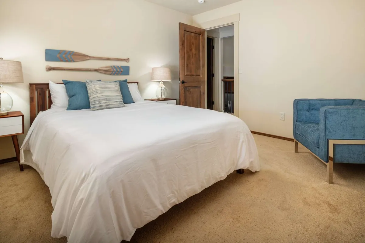 A serene bedroom with white linens, decorative oars mounted on the wall, a blue accent chair by the window, and table lamps providing soft lighting. Natural light filters into the room, creating a calm environment.