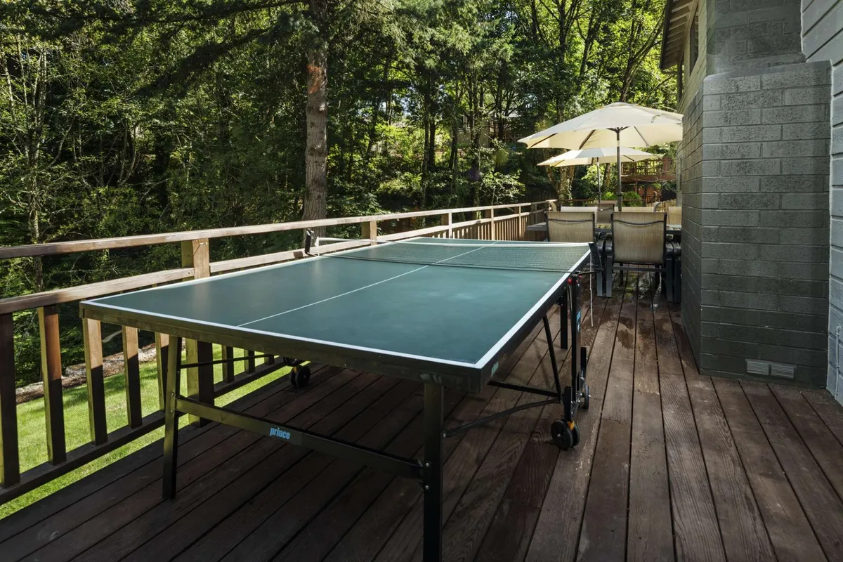 This image captures an expansive outdoor deck featuring a ping pong table, an outdoor dining set, and lush greenery all around. The scene is lively and inviting, with plenty of space for both play and relaxation under Oregon skies.