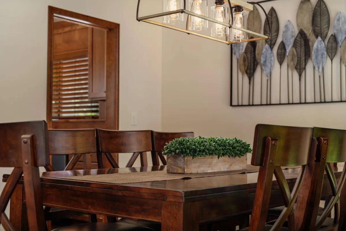 A view of an inviting dining area with a stylish wooden dining table, a hanging light fixture above, decorative wall art in the background, and a lush potted plant. The space feels perfect for both intimate family dinners and festive gatherings.