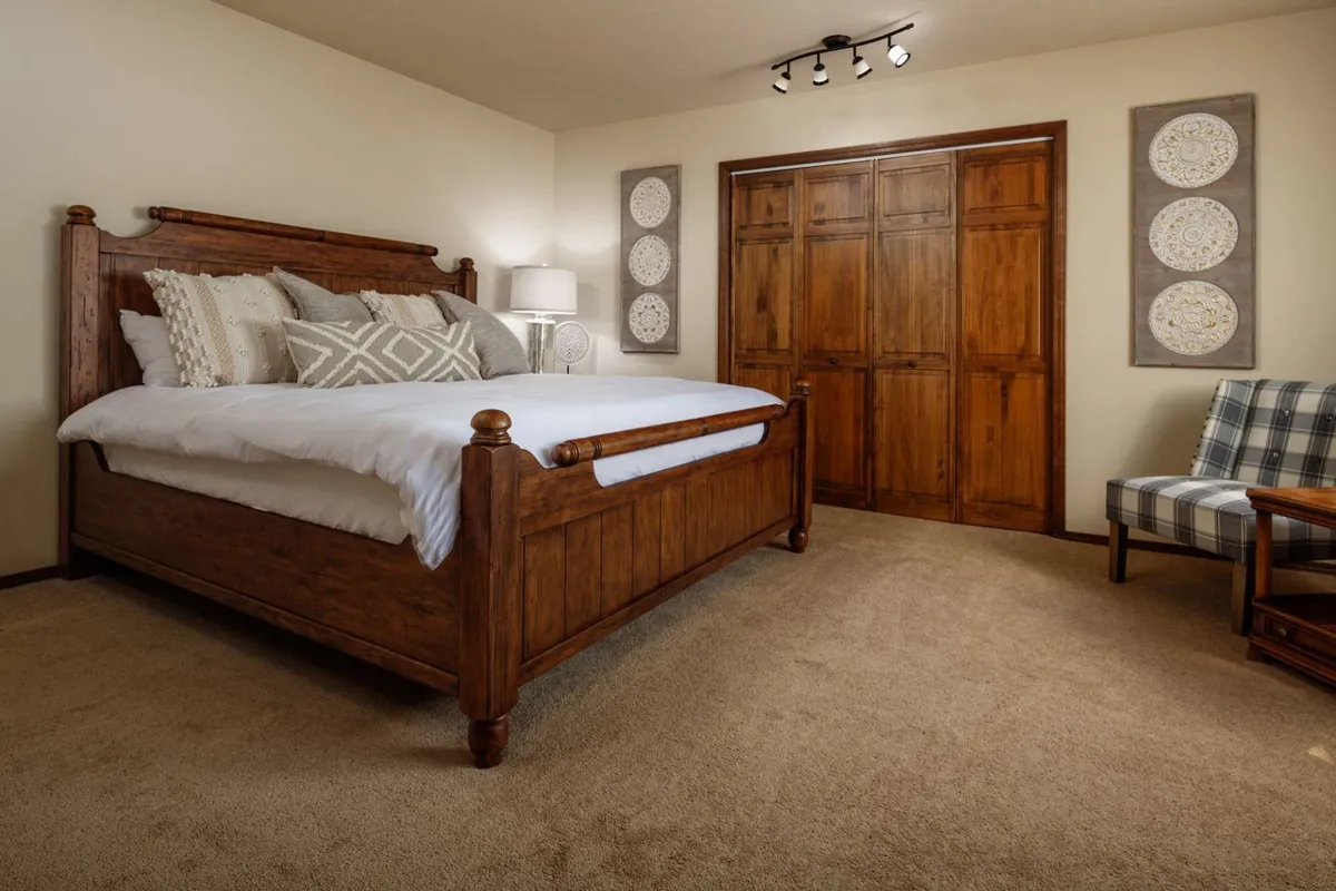 A cozy interior snapshot of the lower-level suite’s bedroom, showcasing a wooden bed frame, decorative pillows, a nightstand with a lamp, storage closet, and an upholstered armchair. The mood is restful and refined, hinting at the suite’s comfort and privacy.