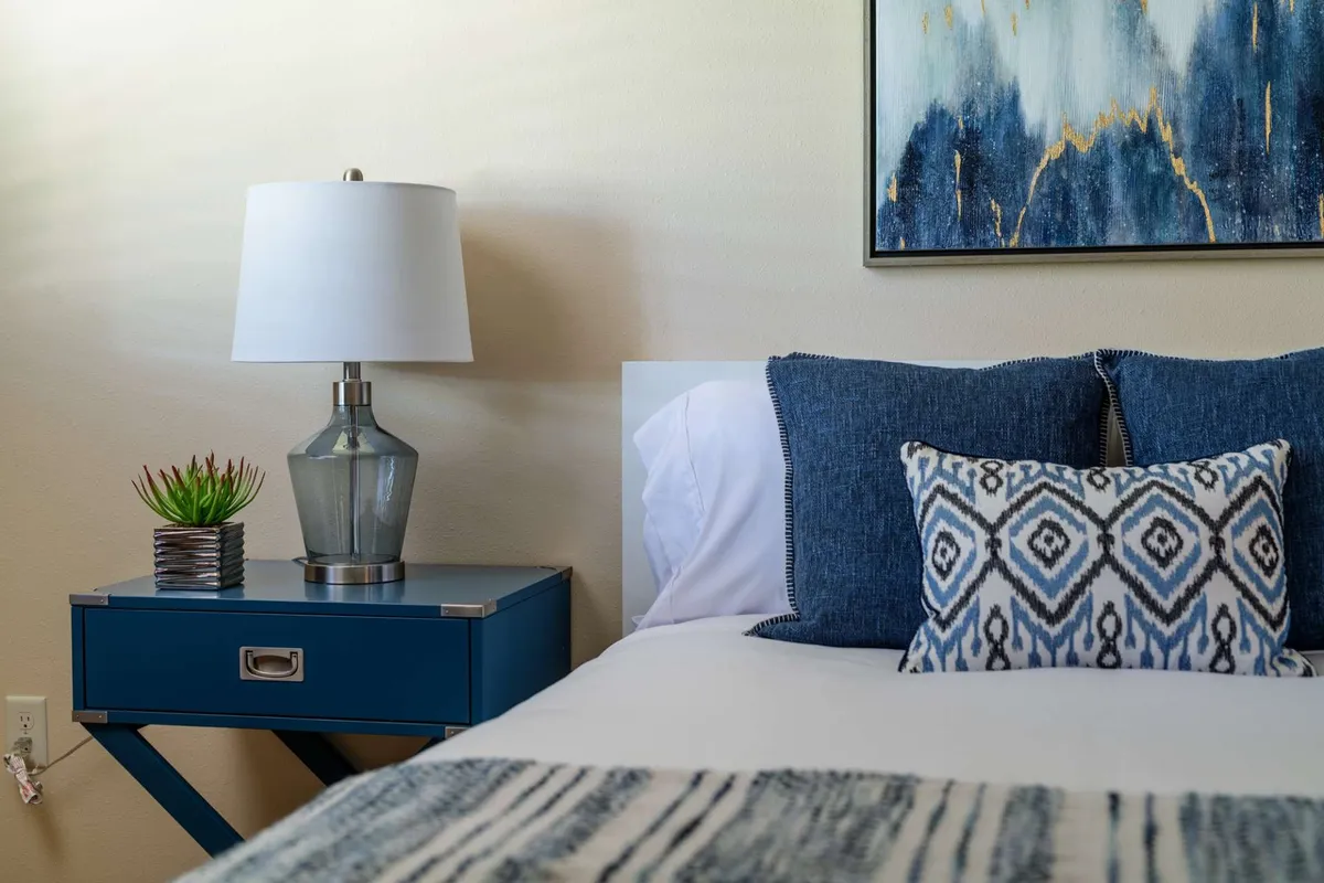 A cozy bedroom bathed in natural light, featuring a comfortable bed with decorative pillows, a modern lamp on the nightstand, and tasteful wall art. The ambiance suggests a restful retreat after an active day.